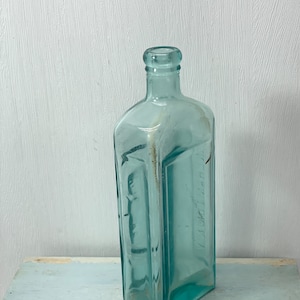 May include: A vintage, rectangular, light blue glass bottle with a narrow neck. The bottle has a slightly faded appearance and appears to be antique. The bottle is sitting on a light blue surface.