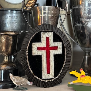 May include: An oval-shaped wall hanging featuring a central cross with a red velvet center and white embroidered border, surrounded by a black pleated fabric frame. The cross is mounted on a black velvet background and edged with a twisted silver cord.