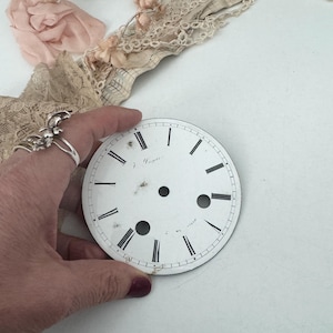 May include: A round white clock dial featuring black Roman numerals, a central hole, and two smaller circular cutouts at the bottom. The dial displays the text J. Dayer in black script and has visible signs of wear and aging on the surface.