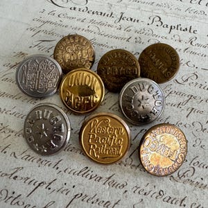 May include: A collection of nine round metal buttons in silver, gold, and bronze tones. Each button features embossed text including New York New Haven and Hartford, Union Pacific, Conductor, Western Pacific Railroad, and Worcester Stry-Co.