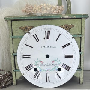 Peut inclure: Un cadran d'horloge rond en émail blanc avec des chiffres romains noirs, un trou central et le texte imprimé MORAND François au Bourg Saint Andéol. Le cadran présente un motif de couronne florale verte et rouge sous le centre.