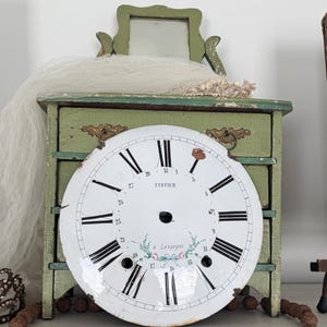 Peut inclure: Un cadran d'horloge blanc rond avec des chiffres romains noirs et des petits chiffres arabes de 1 à 31, portant l'inscription TUFFIER et à Langogne avec un motif floral. Il est placé devant un petit coffre en bois vert vieilli à tiroirs.