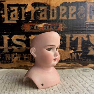 May include: A pale bisque porcelain doll head featuring a smooth, hairless scalp, painted brown eyebrows, open eye sockets, and delicate pink-tinted cheeks with small, painted red lips. A single small circular hole is visible at the base of the neck.