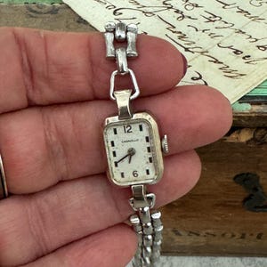 May include: A silver-toned Carvelle wristwatch with a rectangular white face, black hour markers, and black hands. The watch features a metallic link bracelet with a repeating geometric pattern and a small winding crown on the right side.