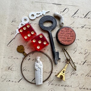 May include: A collection of vintage objects, including red dice, a key, a miniature Eiffel Tower charm, and a small figurine. A small tin reads "FOR RELIEF OF INFLAMED CUTICLE AND HANG NAILS". The items are on a handwritten paper background.