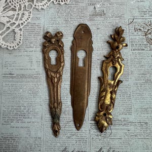 May include: Three antique brass keyhole escutcheon plates arranged vertically. The left plate features a bow and leaf motif, the center plate is a smooth elongated oval, and the right plate displays an ornate floral and leaf design.
