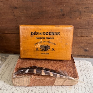 May include: A rectangular wooden box with the black text DÉS A COUDRE, FABRICATION FRANÇAISE, MARQUE DE FABRIQUE, B. D., and DÉPOSÉE, featuring a bee illustration. A brown and black striped feather rests on a weathered, textured book below the box.