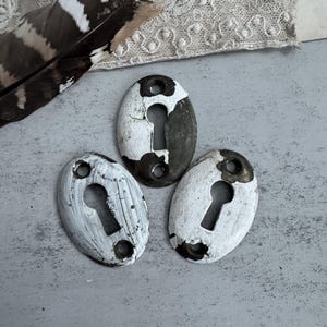 May include: Three oval-shaped metal keyhole escutcheons featuring a distressed white painted finish over a dark metallic base. Each piece has a central keyhole opening and two small circular screw holes at the top and bottom for mounting purposes.