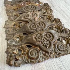 May include: A collection of five antique brass drawer pulls arranged in a vertical stack. Each pull features an ornate, symmetrical scrollwork design with a central leaf motif and two circular screw holes for mounting on furniture surfaces.