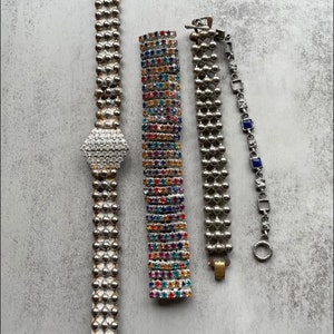 Puede incluir: Cuatro pulseras vintage. Una es un reloj plateado con una esfera en forma de diamante. Otra es una pulsera plateada con pedrería colorida. Las otras dos son pulseras plateadas con varios diseños y cierres.