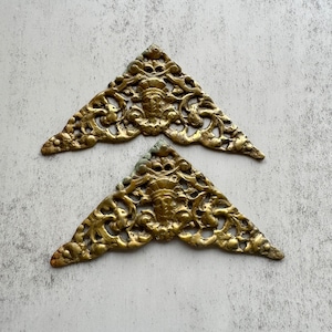 Puede incluir: Dos adornos de esquina antiguos de latón. Cada pieza triangular presenta un diseño ornamentado con una cara central y patrones arremolinados. El latón tiene una pátina cálida y envejecida, lo que sugiere artesanía vintage. Ideal para la restauración de muebles.