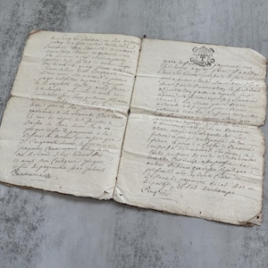May include: An antique, off-white, folded paper document featuring handwritten French cursive script in dark ink. The document includes a small, detailed crest or seal at the top right corner and shows visible signs of age, including frayed edges.