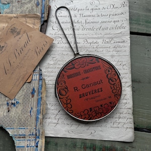 May include: A vintage-style hand mirror with a red circular design featuring the text "BOUCHERIE-CHARCUTERIE" and "R. Canaut BRUYÈRES". The mirror has a silver frame and a black handle. The background includes aged paper and antique ephemera.