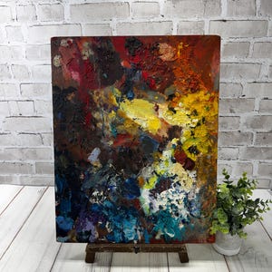 May include: Abstract painting with a rich palette of colours, including red, yellow, blue, and brown. The artwork is displayed on a small wooden easel, highlighting the textured brushstrokes and artistic style. A unique piece of art.