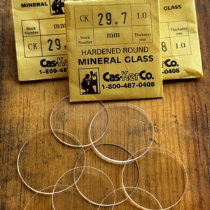 Op de afbeelding: Transparante, ronde minerale glazen horlogekristallen met een dikte van 1,0 mm. De afbeelding toont ook gele verpakkingen met de tekst "HARDENED ROUND MINERAL GLASS" en het Cas-KerCo.-logo.