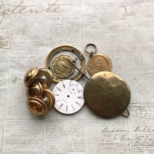 May include: A collection of vintage watch parts including a white circular clock face with black Roman numerals, a brass pendulum, a brass ring, a small circular medal with embossed text, and a cluster of four smooth, round brass buttons.