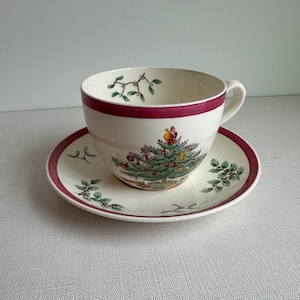 May include: A cream-colored ceramic teacup and saucer set featuring a festive Christmas tree graphic with ornaments and a figure at the top. The rim of the cup and the edge of the saucer are accented with a solid dark red band and green holly leaves.