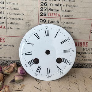 Può includere: Un quadrante d'orologio rotondo in smalto bianco con numeri romani neri per le ore e numeri arabi neri per i minuti a intervalli di cinque. Il quadrante presenta un foro centrale e due aperture circolari più piccole posizionate in basso.