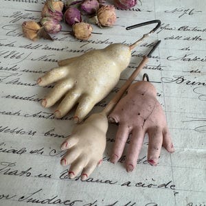 May include: Three vintage doll hands made of composition or ceramic material. Two hands are a pale cream color with painted red fingernails, and one hand is a dusty pink color with visible cracks and painted red fingernails, attached to metal hooks.