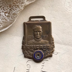 May include: A bronze-toned metal badge featuring a relief portrait of General Pershing with the text 'GENERAL PERSHING' and '1939'. Below the portrait is the word 'CHICAGO' and a small circular emblem with blue enamel reading 'AMERICAN LEGION'.
