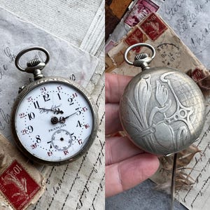 May include: Antique silver pocket watch with a white face, black numerals, and red accents. The watch face reads "Regulateur Francais". The back of the watch features an engraved floral design. The watch is displayed on a background of vintage papers and stamps.
