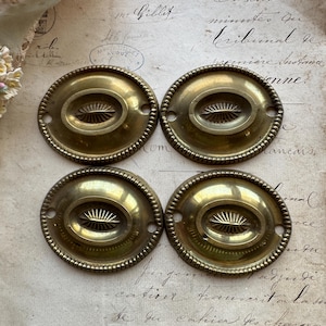 May include: Four antique brass oval drawer pulls. Each pull has a central oval design with a sunburst pattern and a beaded border. The pulls are arranged on a vintage paper background.