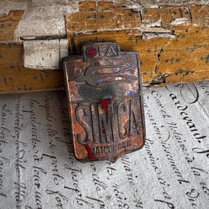 Può includere: Un'insegna per auto SIMCA vintage con un disegno di uccello e le scritte "SIMCA" e "NANTERRE". L'emblema è nei toni del rame, del rosso e del blu. Lo sfondo è una superficie strutturata con scritte a mano.