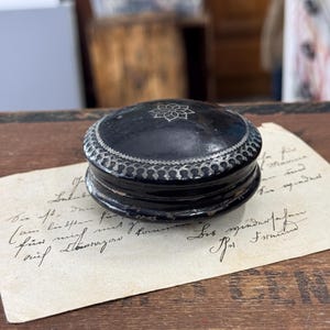 Puede incluir: Pequeña caja redonda negra con un diseño floral plateado en la tapa y un borde plateado decorativo. La caja descansa sobre un trozo de papel envejecido con texto manuscrito. La caja es probablemente vintage o antigua.