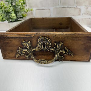 May include: A vintage wooden drawer with a decorative brass handle. The drawer is stained a dark brown colour and features ornate floral detailing on the handle. The drawer is empty and sits on a white surface.