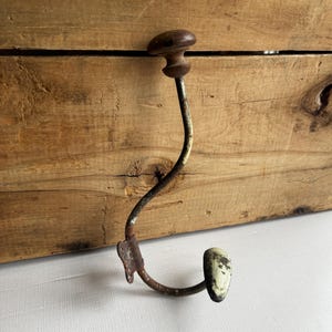 Puede incluir: Un gancho de metal antiguo con un diseño curvo. El gancho tiene un pomo de madera marrón oscuro en la parte superior y un cuerpo de metal oxidado y curvado. El extremo del gancho está pintado con un color blanquecino descolorido. El gancho está montado sobre una superficie de madera.