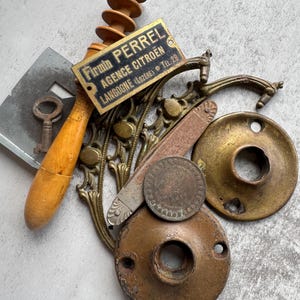Op de afbeelding: Een verzameling vintage ijzerwaren, waaronder een houten honingdipper, een metalen sleutel, een rechthoekig messing bordje met de tekst Firmin Perrel Agence Citroën Langogne (Lozère) Tel 28, een decoratieve messing beugel, een handvat en twee ronde platen.