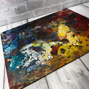 May include: A rectangular wooden artist's palette with a colourful mix of oil paint. The palette has a textured surface with bold strokes of yellow, blue, red, and white paint, creating an abstract design. The palette is set against a white wooden background.