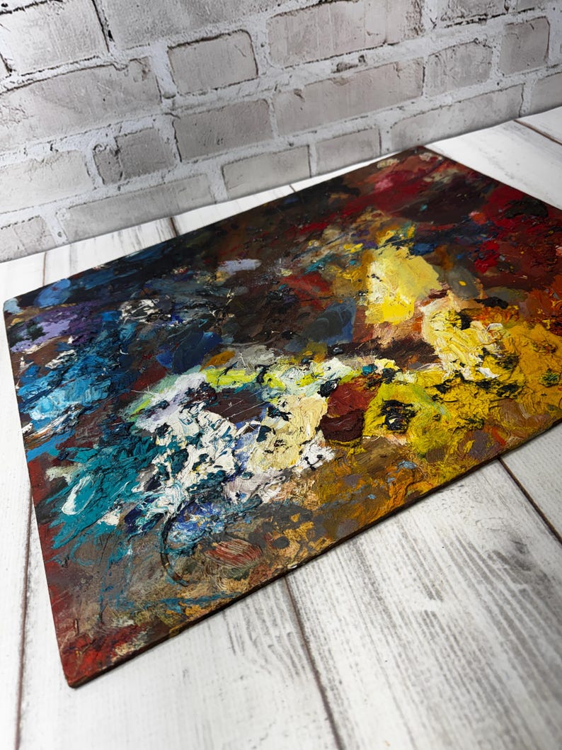 May include: A rectangular wooden artist's palette with a colourful mix of oil paint. The palette has a textured surface with bold strokes of yellow, blue, red, and white paint, creating an abstract design. The palette is set against a white wooden background.
