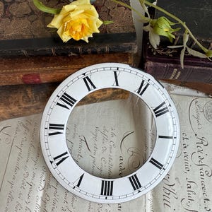 May include: A circular white clock dial featuring black Roman numerals. The dial has a hollow center and a smooth, glossy finish. The black markings are clearly defined against the white background, forming a classic ring-shaped clock face.