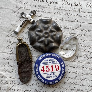 Puede incluir: Un surtido de objetos antiguos que incluyen un colgante de cruz blanca, un objeto metálico en forma de flor, un cristal transparente, un pequeño monedero de cuero y un botón de feria azul y blanco con el número 4519.