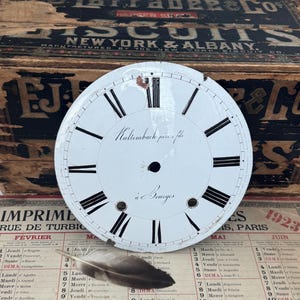 Peut inclure: Un cadran d'horloge circulaire en émail blanc avec des chiffres romains noirs, un trou central et deux petites ouvertures circulaires en bas. Le texte "Kaltembach père & fils" et "à Bourges" est imprimé en écriture noire au centre.