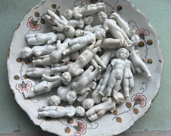 Może przedstawiać: Kolekcja małych, białych, nieszkliwionych figurek lalek z porcelany ułożonych w okrągłym ceramicznym talerzu z falistym brzegiem, ozdobionym powtarzającym się wzorem różowych kwiatów, zielonych liści i małych pomarańczowych akcentów na obrzeżu.