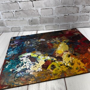 May include: A rectangular artist's palette displaying an abstract painting with bold brushstrokes. The artwork uses a palette of blues, yellows, reds, and browns. The palette sits on a white wooden surface, with a grey brick wall behind it.