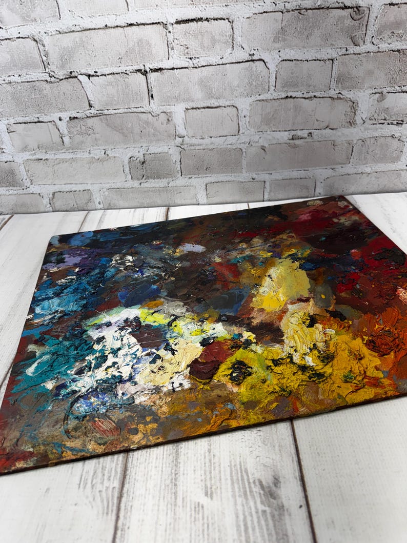 May include: A rectangular artist's palette displaying an abstract painting with bold brushstrokes. The artwork uses a palette of blues, yellows, reds, and browns. The palette sits on a white wooden surface, with a grey brick wall behind it.