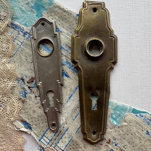 Może przedstawiać: Dwie zabytkowe płytki do klamek. Jedna jest posrebrzana, druga mosiężna. Obie mają otwór na klucz i okrągły otwór na klamkę. Srebrna płytka ma wzór Art Deco. Mosiężna płytka ma bardziej ozdobny, zaokrąglony kształt.