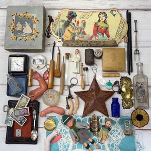 Puede incluir: Una colección de objetos antiguos, que incluye un reloj, muñecas, un adorno en forma de estrella y varias herramientas. Los objetos están dispuestos sobre una superficie de madera blanca, con una variedad de colores y texturas.