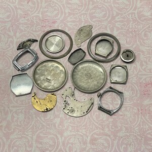 May include: A collection of vintage watch parts, including silver-toned cases, rings, and internal mechanisms. The components are scattered on a pink floral patterned surface, showcasing the intricate details of horology.