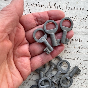 May include: A collection of antique metal keys, some held in a hand, others scattered below. The keys have a vintage, worn appearance, with circular keyholes and detailed teeth. The background features faded script.
