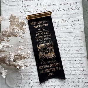 May include: A black ribbon with gold lettering and a gold-coloured metal clasp. The ribbon reads "NEW HAMPSHIRE BATTALION K.T. PILGRIM COMMANDERY LACONIA" and "Pilgrimage to TRIENNIAL CONCLAVE WASHINGTON D.C. 1889."