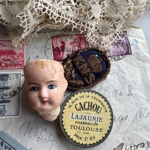 May include: A vintage doll head with blue eyes and a small, round, yellow and black tin of Cachou Lajaunie. A navy blue and brown decorative pin and antique stamps are also visible.
