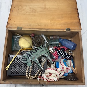 Puede incluir: Una caja de madera abierta llena de objetos antiguos. Contenido: botellas de vidrio, un rallador de metal, un collar de perlas, un caballo de metal con texto "SAKATA Visual Communication" y varios otros objetos antiguos.