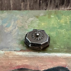 May include: A small, octagonal metal box with a dark, weathered finish. The lid features a central, raised silver-toned sunburst or floral emblem with a small, rounded knob at its center. The edges of the lid are defined by a thin, bright silver border.