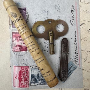 May include: A vintage Stein's Lining Color Stick in a cylindrical paper tube, a brass clock winding key with a butterfly-shaped handle, and a dark brown rectangular wax seal stamp, all resting on a vintage envelope with postage stamps and script.