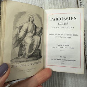 Puede incluir: Un libro antiguo abierto con una ilustración detallada de San Juan Evangelista en la página izquierda. La página derecha muestra texto en francés, incluyendo "PAROISSIEN ROMAIN" y "PARTIE D'HIVER". La portada del libro es visible.