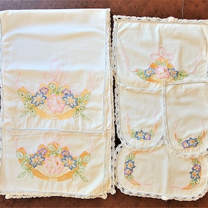 May include: A cream-colored embroidered table runner and matching placemats. The floral design features blue, pink, and yellow flowers with green leaves. The edges are trimmed with a delicate lace.
